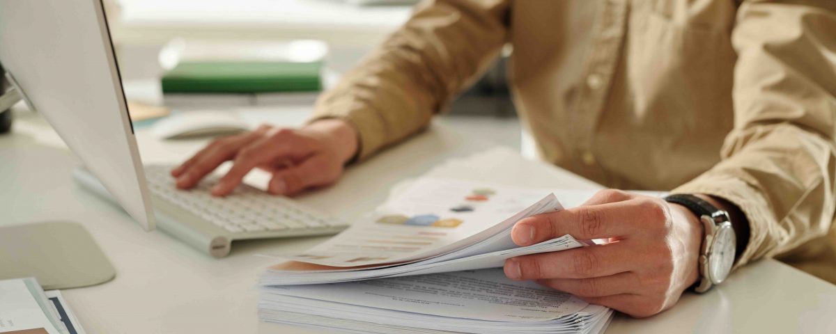 Focus on hand of young financial director of marketing company taking some documents from stack of papers while working by desk