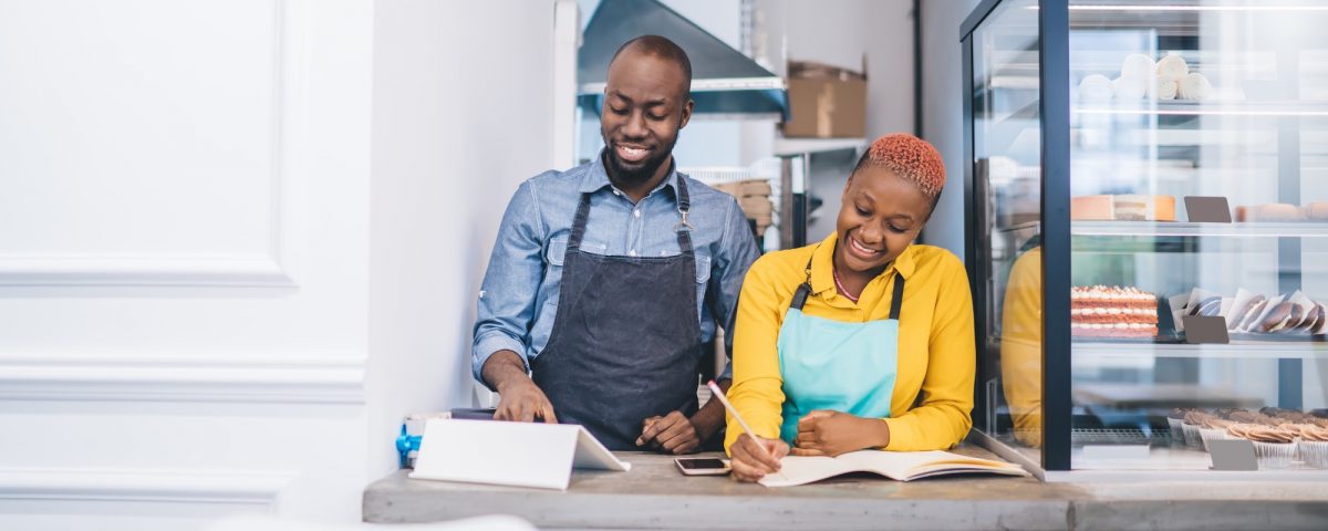 Smiling African American cooks in yellow and gray shirts and blue and black aprons checking information in tablet and writing to notebook behind counter of pastry shop